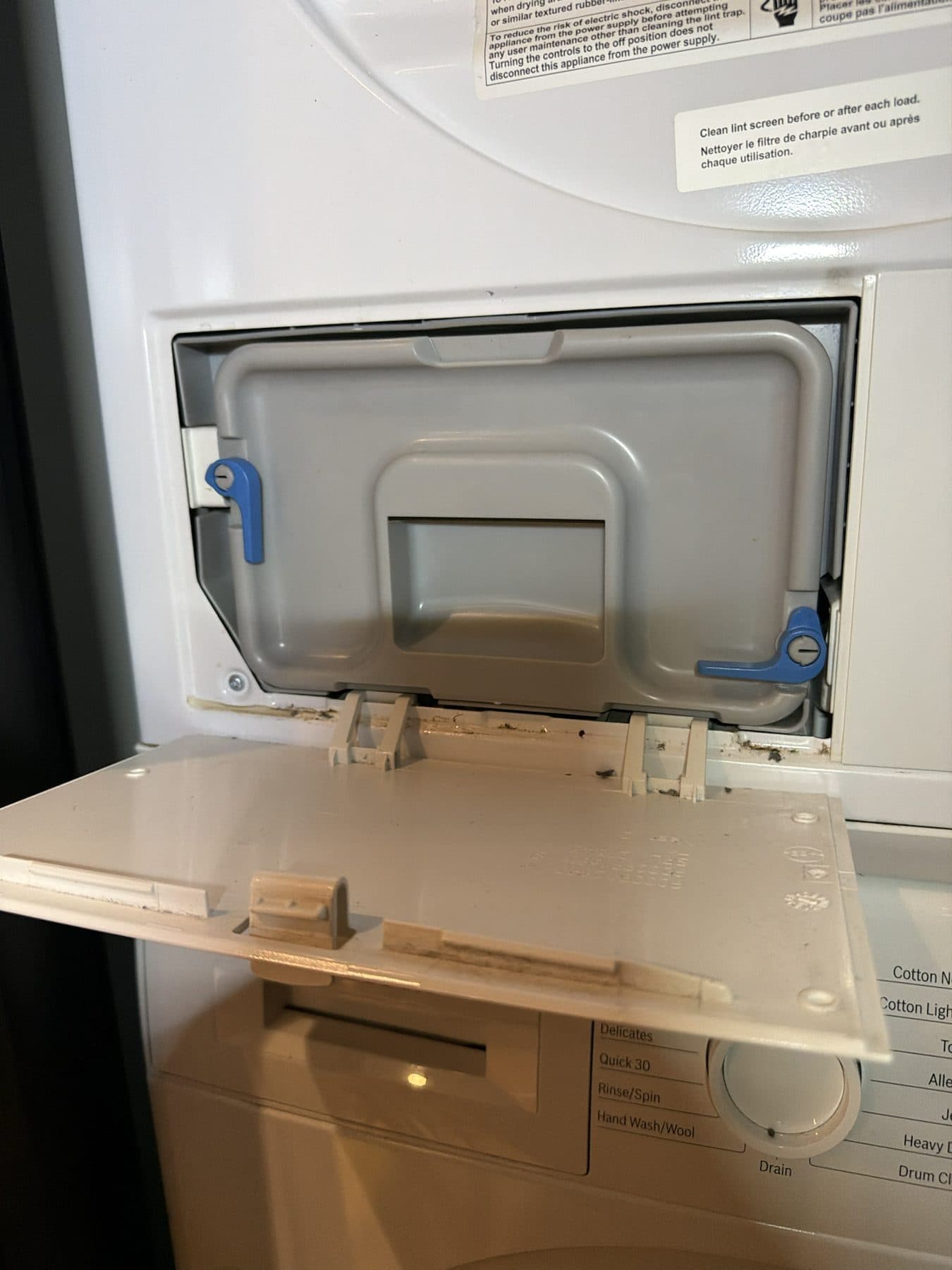 The bottom front flap of a ventless dryer folded open, showing the grey condenser cover held in place by two blue release levers