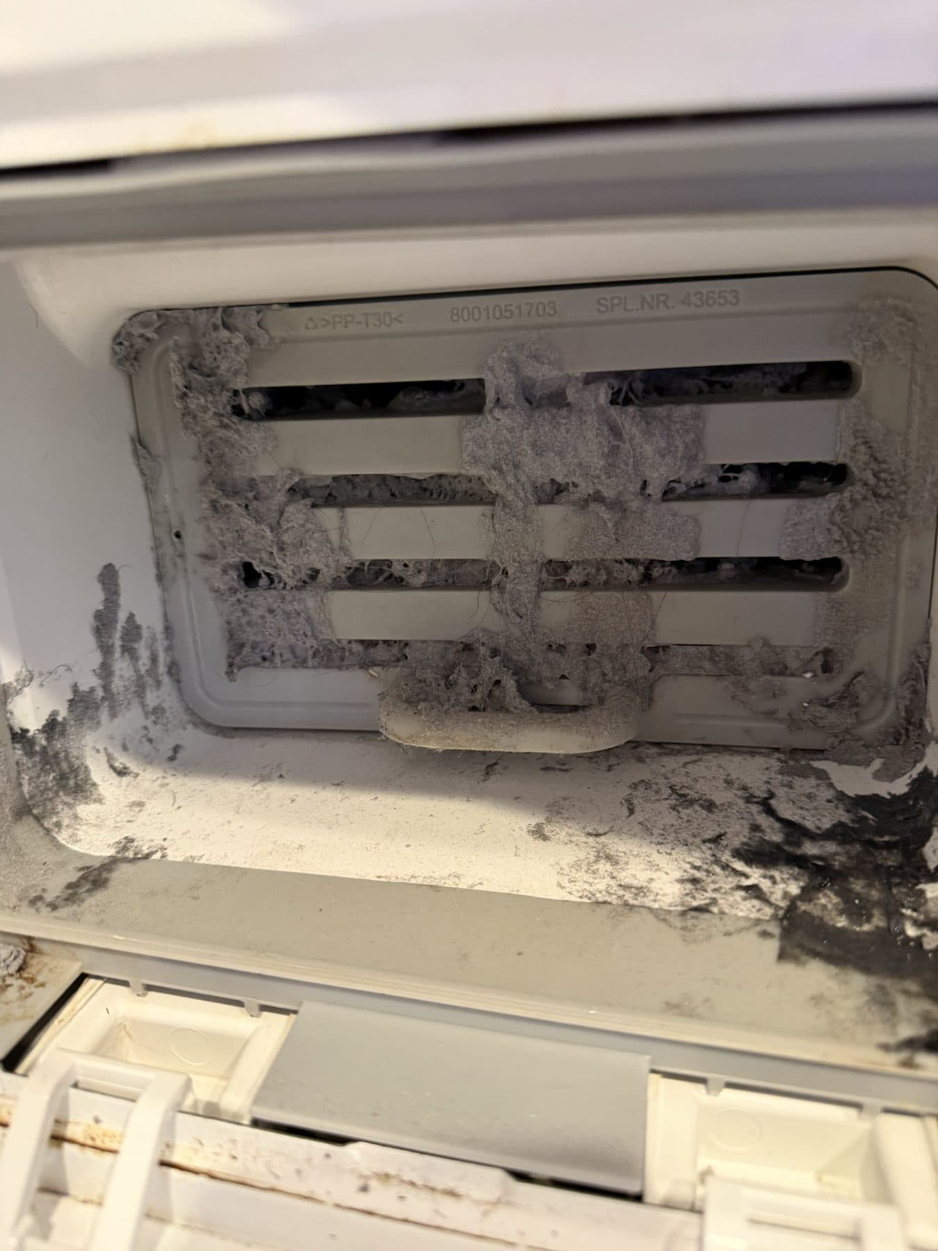 The condenser compartment of a ventless dryer packed with grey lint and dark buildup across the condenser grille and the floor of the compartment
