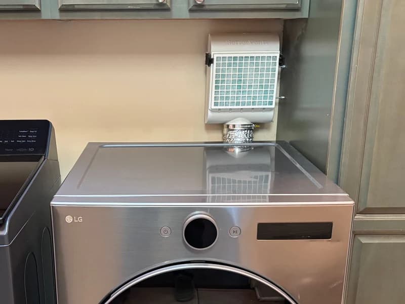 A white BetterVent indoor dryer vent mounted on the wall above a stainless front-load dryer, with its wide filter pad facing the room and a short flexible duct running down to the dryer