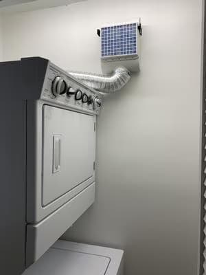 A BetterVent indoor dryer vent mounted high on the wall beside a white stacked washer-dryer unit, connected by a curved flexible duct