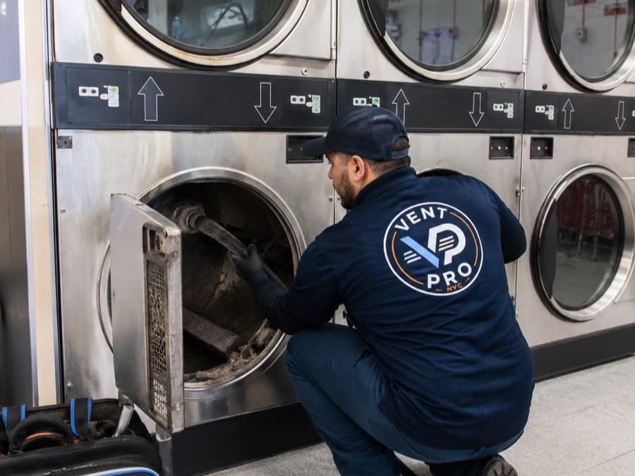 Vent Pro NYC technician vacuuming lint from the open lint compartment of a stacked commercial dryer in a laundromat