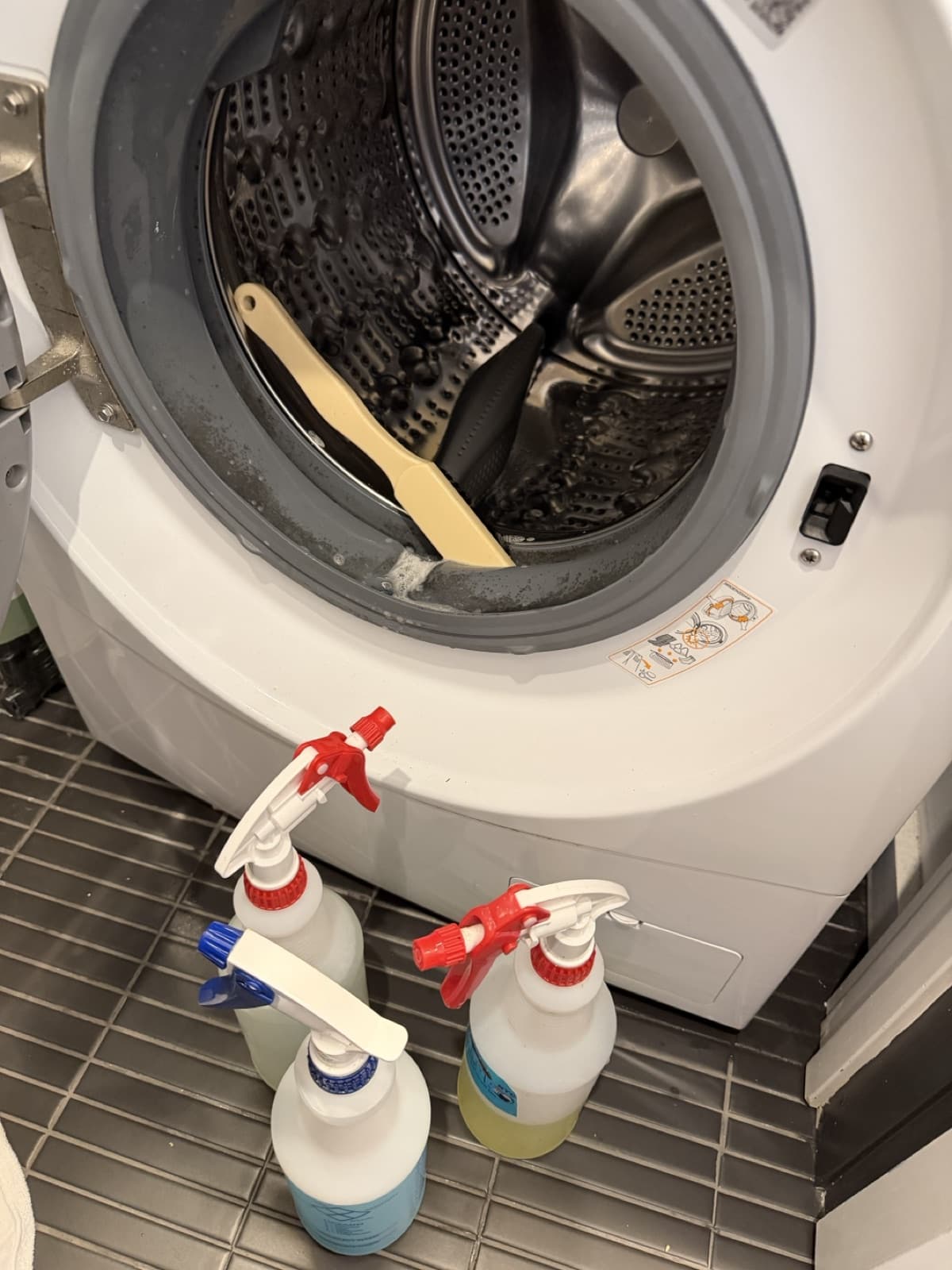 A scrub brush resting in the sudsy door-gasket channel of a front-load washer mid-treatment, spray bottles on the floor below