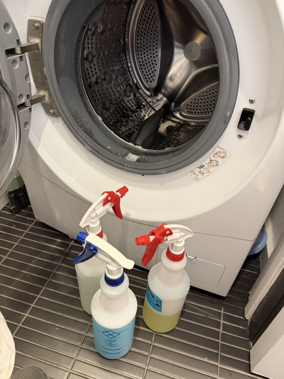 Three unlabeled spray bottles of Vent Pro NYC's own gasket-cleaning solutions lined up on the floor in front of an open front-load washer