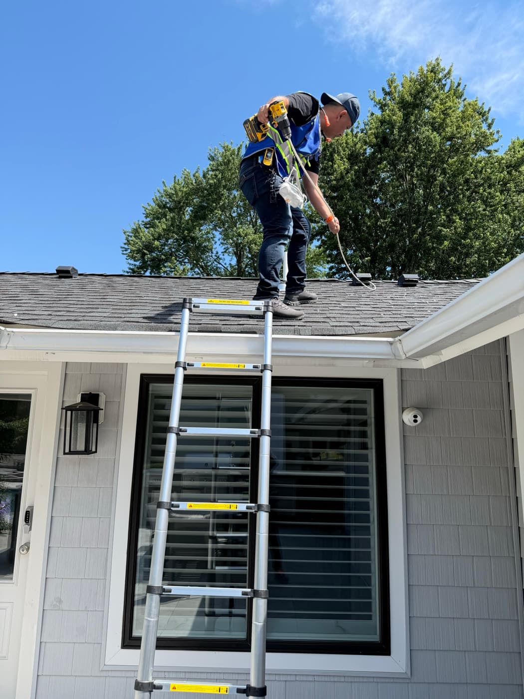A Vent Pro NYC technician on the roof of a home, using a drill-driven brush to clear a roof dryer vent, telescoping ladder extended to the gutter