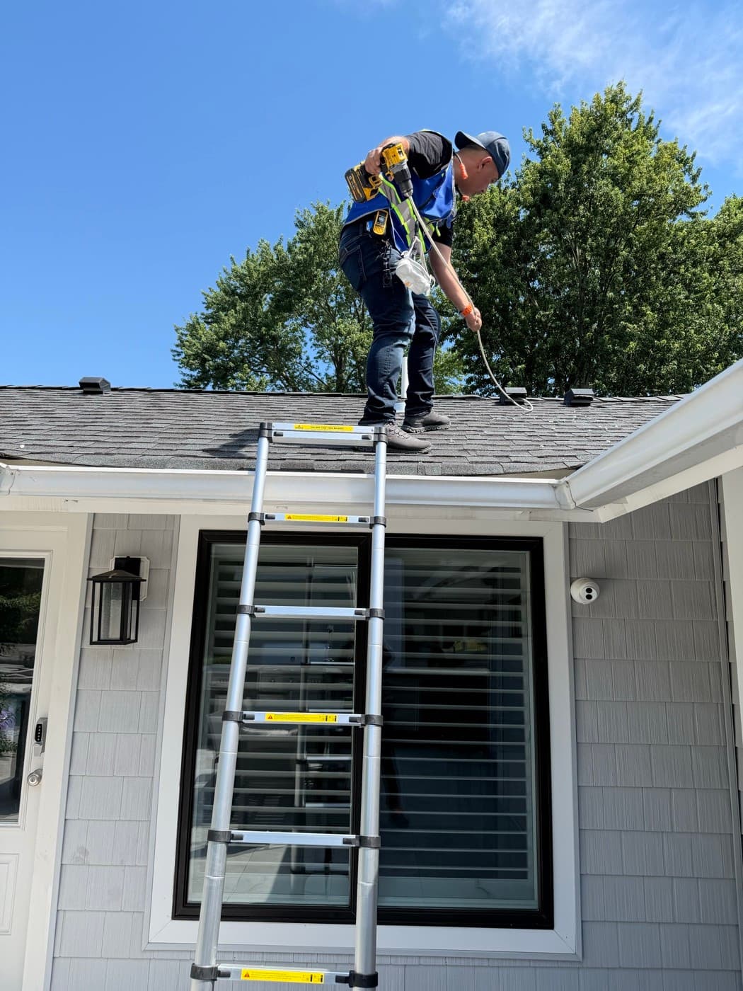 A Vent Pro NYC technician on the roof of a home, using a drill-driven brush to clear a roof dryer vent, telescoping ladder extended to the gutter